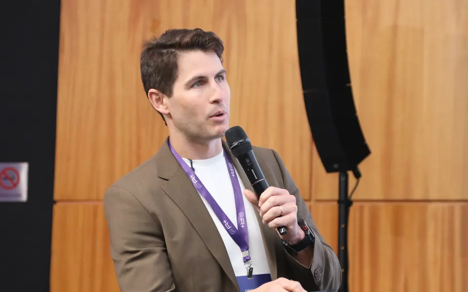 Professor falando ao microfone no palco de um evento da R+