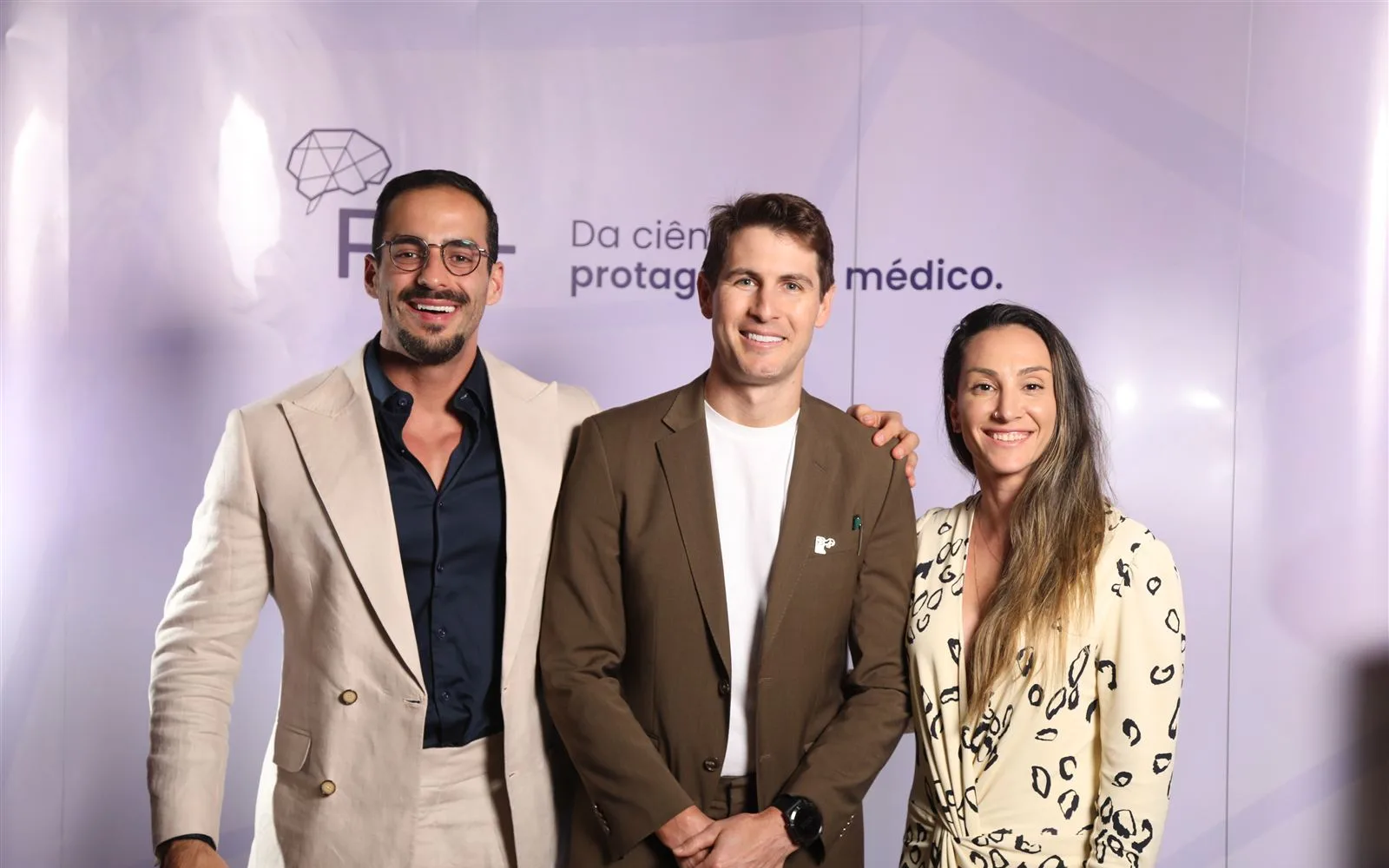 Dr. Thomáz Baêsso, Dr. Guilherme Renke e Dra. Thaís Fonseca juntos, sorrindo, em frente ao painel do R+ num congresso
