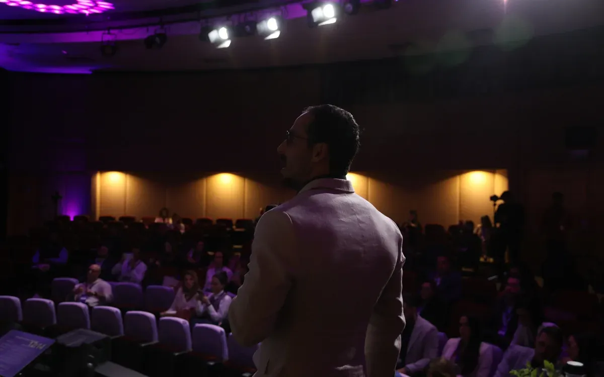 Palestrante no palco, de costas, diante do auditório cheio num evento da R+