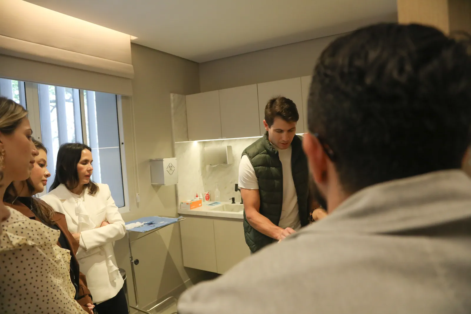 Sala clínica com pia e bandeja de material, médicos observando o mentor